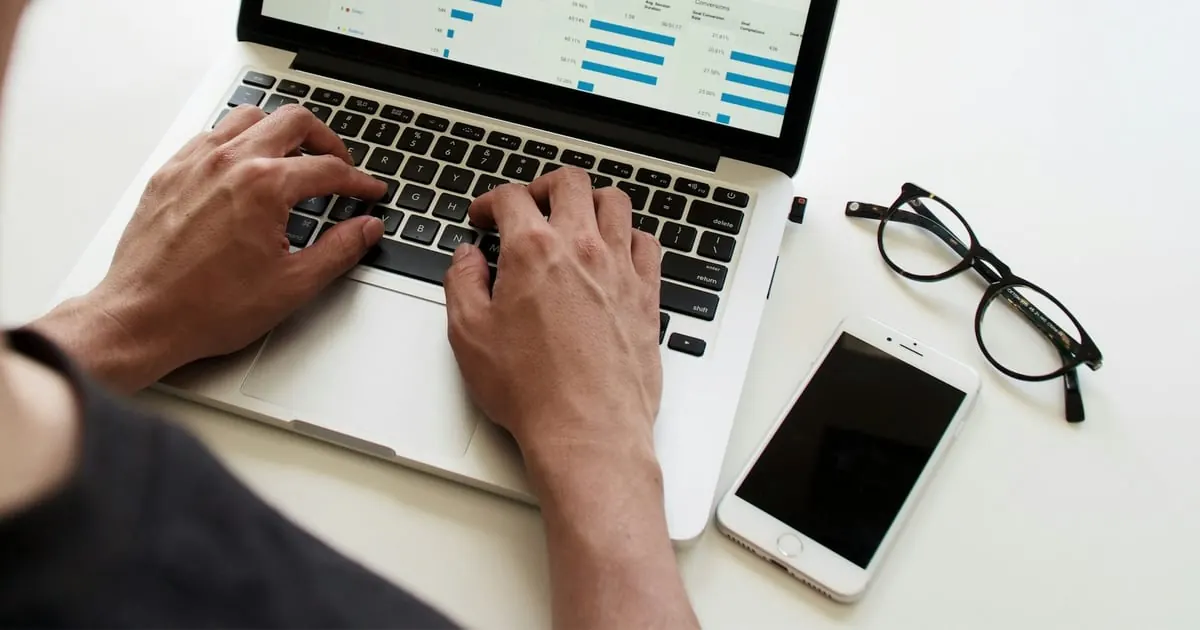 Dłonie na klawiaturze laptopa, obok okulary i telefon